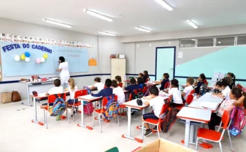 Sala de aula infantil em Florianópolis, com crianças escrevendo em cadernos enquanto a professora escreve no quadro, representando a educação na rede municipal.