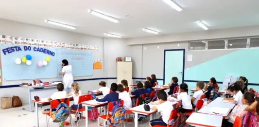 Sala de aula infantil em Florianópolis, com crianças escrevendo em cadernos enquanto a professora escreve no quadro, representando a educação na rede municipal.