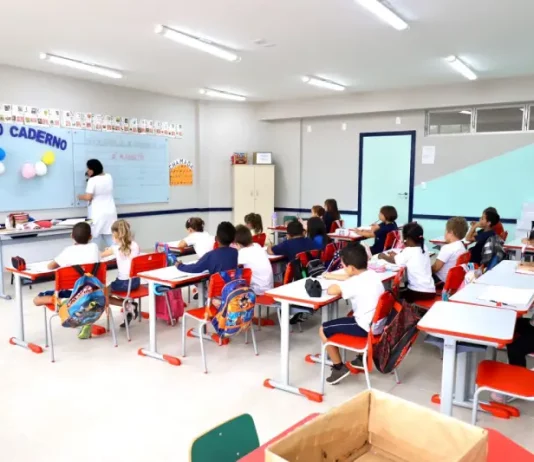 Sala de aula infantil em Florianópolis, com crianças escrevendo em cadernos enquanto a professora escreve no quadro, representando a educação na rede municipal.