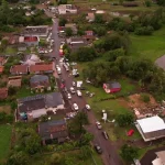 Vista aérea mostra parte de Dionísio Cerqueira após a passagem de tornados, com casas destelhadas, destroços espalhados e equipes atuando nas ruas para avaliar os danos e prestar atendimento à população. A imagem ilustra a dimensão dos estragos causados pelo fenômeno climático.