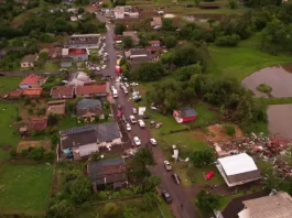 Vista aérea mostra parte de Dionísio Cerqueira após a passagem de tornados, com casas destelhadas, destroços espalhados e equipes atuando nas ruas para avaliar os danos e prestar atendimento à população. A imagem ilustra a dimensão dos estragos causados pelo fenômeno climático.