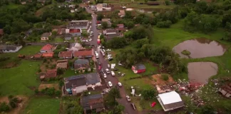 Vista aérea mostra parte de Dionísio Cerqueira após a passagem de tornados, com casas destelhadas, destroços espalhados e equipes atuando nas ruas para avaliar os danos e prestar atendimento à população. A imagem ilustra a dimensão dos estragos causados pelo fenômeno climático.