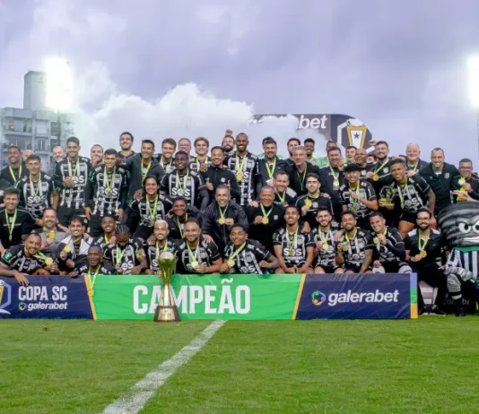 Imagem do elenco e comissão técnica do Figueirense reunidos no gramado do Estádio Orlando Scarpelli para registrar a celebração do título da Copa SC, destacando o troféu ao centro e a faixa de campeão exibida na frente do grupo.