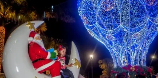 A imagem mostra uma cena natalina usada para divulgar o Natal Iluminado de São José. Um Papai Noel decorativo está sentado em uma lua gigante ao lado de uma criança, enquanto ao fundo aparece uma grande estrutura iluminada em azul em formato de balão e famílias aproveitam o espaço. A foto reforça o clima festivo e a proposta de oferecer atrações para as famílias durante o evento.