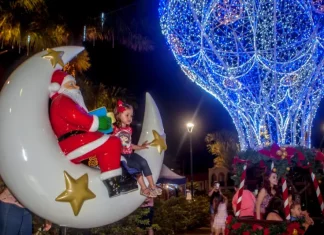 A imagem mostra uma cena natalina usada para divulgar o Natal Iluminado de São José. Um Papai Noel decorativo está sentado em uma lua gigante ao lado de uma criança, enquanto ao fundo aparece uma grande estrutura iluminada em azul em formato de balão e famílias aproveitam o espaço. A foto reforça o clima festivo e a proposta de oferecer atrações para as famílias durante o evento.