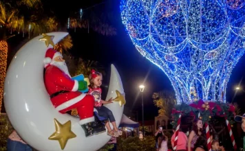 A imagem mostra uma cena natalina usada para divulgar o Natal Iluminado de São José. Um Papai Noel decorativo está sentado em uma lua gigante ao lado de uma criança, enquanto ao fundo aparece uma grande estrutura iluminada em azul em formato de balão e famílias aproveitam o espaço. A foto reforça o clima festivo e a proposta de oferecer atrações para as famílias durante o evento.