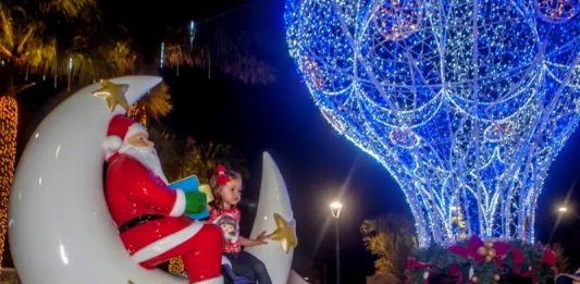 São José lança o Natal Iluminado e estreia a Vila Mágica do Natal Luzes da Esperança A imagem mostra uma cena natalina usada para divulgar o Natal Iluminado de São José. Um Papai Noel decorativo está sentado em uma lua gigante ao lado de uma criança, enquanto ao fundo aparece uma grande estrutura iluminada em azul em formato de balão e famílias aproveitam o espaço. A foto reforça o clima festivo e a proposta de oferecer atrações para as famílias durante o evento.
