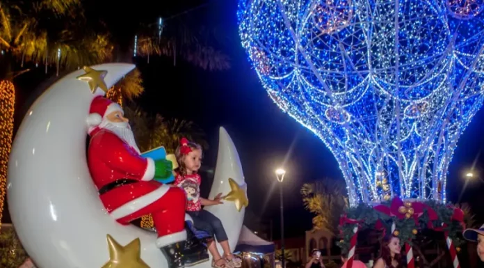 A imagem mostra uma cena natalina usada para divulgar o Natal Iluminado de São José. Um Papai Noel decorativo está sentado em uma lua gigante ao lado de uma criança, enquanto ao fundo aparece uma grande estrutura iluminada em azul em formato de balão e famílias aproveitam o espaço. A foto reforça o clima festivo e a proposta de oferecer atrações para as famílias durante o evento.