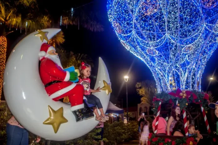Natal SJ A imagem mostra uma cena natalina usada para divulgar o Natal Iluminado de São José. Um Papai Noel decorativo está sentado em uma lua gigante ao lado de uma criança, enquanto ao fundo aparece uma grande estrutura iluminada em azul em formato de balão e famílias aproveitam o espaço. A foto reforça o clima festivo e a proposta de oferecer atrações para as famílias durante o evento.