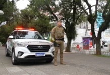 Policial militar uniformizado em frente a viatura, em praça pública, representando a aplicação de multa e a fiscalização do uso de drogas em espaços públicos.