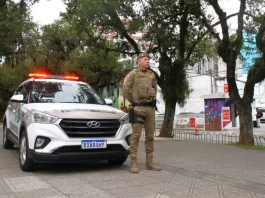 Policial militar uniformizado em frente a viatura, em praça pública, representando a aplicação de multa e a fiscalização do uso de drogas em espaços públicos.