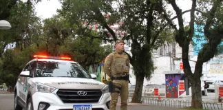 Policial militar uniformizado em frente a viatura, em praça pública, representando a aplicação de multa e a fiscalização do uso de drogas em espaços públicos.