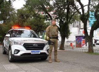 Policial militar uniformizado em frente a viatura, em praça pública, representando a aplicação de multa e a fiscalização do uso de drogas em espaços públicos.