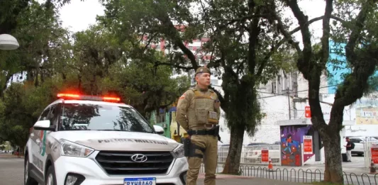 Policial militar uniformizado em frente a viatura, em praça pública, representando a aplicação de multa e a fiscalização do uso de drogas em espaços públicos.
