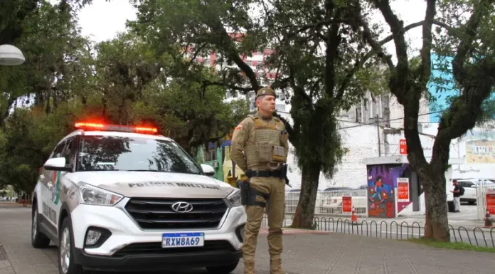 Policial militar uniformizado em frente a viatura, em praça pública, representando a aplicação de multa e a fiscalização do uso de drogas em espaços públicos.
