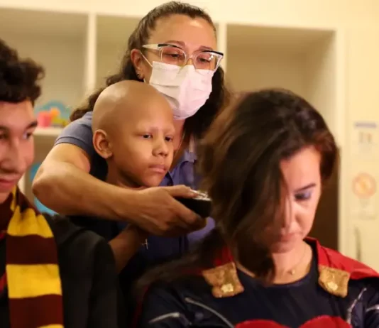 Super-heróis têm cabelo raspado em ação pelo diagnóstico precoce do câncer infantil Criança paciente com câncer infantil raspando o cabelo de um super-herói, simbolizando solidariedade e a campanha de diagnóstico precoce.