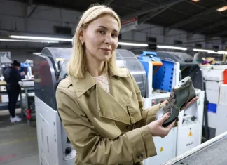 A imagem mostra a empresária Suzana Santos em uma fábrica de calçados, segurando um modelo de sapato para representar a presença e a liderança das mulheres no empreendedorismo catarinense.