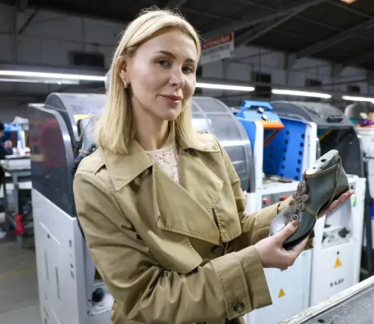 A imagem mostra a empresária Suzana Santos em uma fábrica de calçados, segurando um modelo de sapato para representar a presença e a liderança das mulheres no empreendedorismo catarinense.