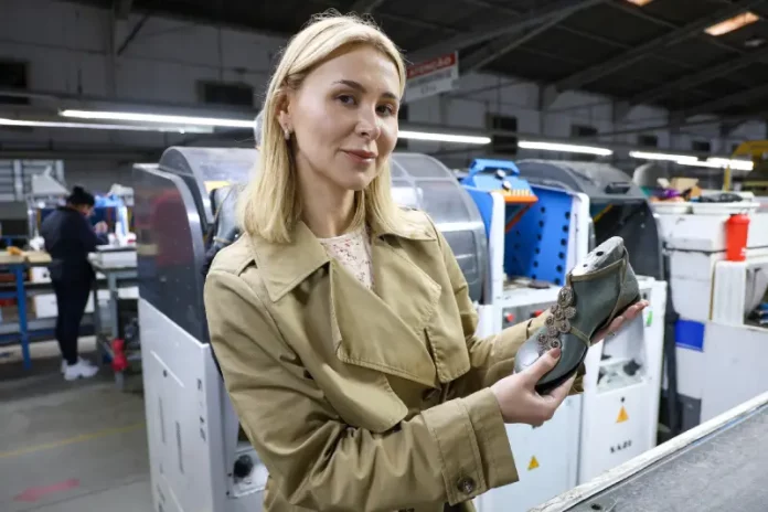 A imagem mostra a empresária Suzana Santos em uma fábrica de calçados, segurando um modelo de sapato para representar a presença e a liderança das mulheres no empreendedorismo catarinense.