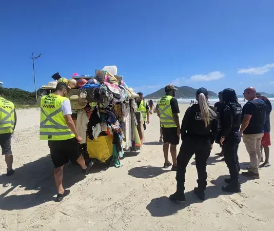 Agentes da Prefeitura e da Polícia Militar abordam um carrinho carregado de produtos irregulares nas praias para ilustrar a fiscalização contra o comércio ambulante ilegal em Florianópolis.