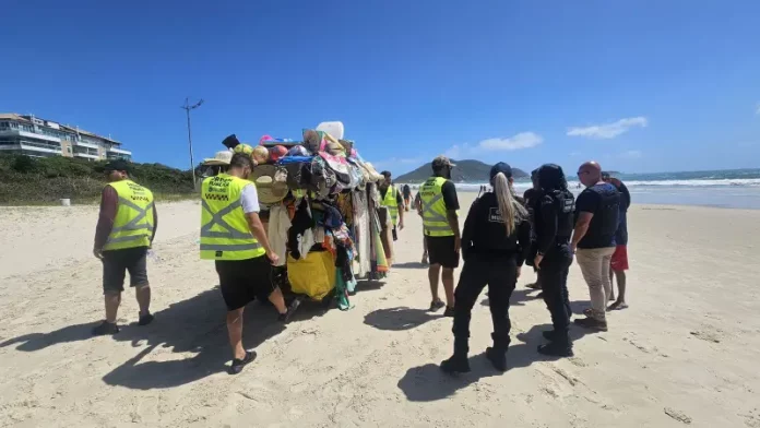 Agentes da Prefeitura e da Polícia Militar abordam um carrinho carregado de produtos irregulares nas praias para ilustrar a fiscalização contra o comércio ambulante ilegal em Florianópolis.