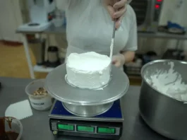 Pessoa realiza trabalho de confeitaria ao aplicar cobertura em um bolo na cozinha de uma unidade prisional, durante atividade de capacitação profissional.