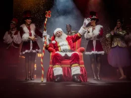 Florianópolis divulga programação completa do Natal 2025 – 100 anos de Luz Foto do Papai Noel sentado em sua poltrona na nova Casa do Papai Noel, rodeado de suas assistentes, com decoração natalina ao redor, transmitindo o clima festivo e acolhedor do Natal em Florianópolis.