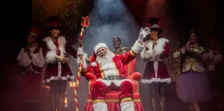 Foto do Papai Noel sentado em sua poltrona na nova Casa do Papai Noel, rodeado de suas assistentes, com decoração natalina ao redor, transmitindo o clima festivo e acolhedor do Natal em Florianópolis.