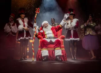Foto do Papai Noel sentado em sua poltrona na nova Casa do Papai Noel, rodeado de suas assistentes, com decoração natalina ao redor, transmitindo o clima festivo e acolhedor do Natal em Florianópolis.