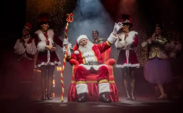 Foto do Papai Noel sentado em sua poltrona na nova Casa do Papai Noel, rodeado de suas assistentes, com decoração natalina ao redor, transmitindo o clima festivo e acolhedor do Natal em Florianópolis.