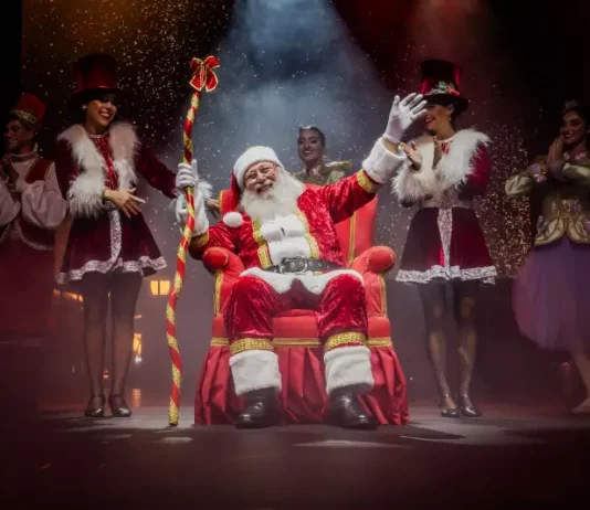 Foto do Papai Noel sentado em sua poltrona na nova Casa do Papai Noel, rodeado de suas assistentes, com decoração natalina ao redor, transmitindo o clima festivo e acolhedor do Natal em Florianópolis.