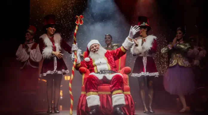 Foto do Papai Noel sentado em sua poltrona na nova Casa do Papai Noel, rodeado de suas assistentes, com decoração natalina ao redor, transmitindo o clima festivo e acolhedor do Natal em Florianópolis.