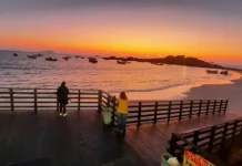 Pessoas observam o pôr do sol em um deque à beira-mar, com barcos ancorados e o céu alaranjado iluminando a praia.