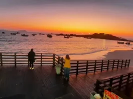 Pessoas observam o pôr do sol em um deque à beira-mar, com barcos ancorados e o céu alaranjado iluminando a praia.