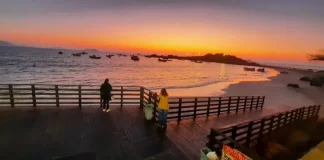 Pessoas observam o pôr do sol em um deque à beira-mar, com barcos ancorados e o céu alaranjado iluminando a praia.