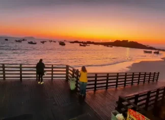 Pessoas observam o pôr do sol em um deque à beira-mar, com barcos ancorados e o céu alaranjado iluminando a praia.