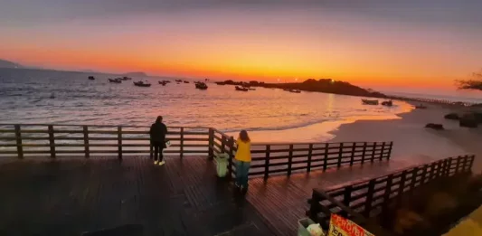 Pessoas observam o pôr do sol em um deque à beira-mar, com barcos ancorados e o céu alaranjado iluminando a praia.