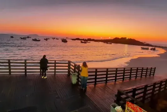 Pessoas observam o pôr do sol em um deque à beira-mar, com barcos ancorados e o céu alaranjado iluminando a praia.