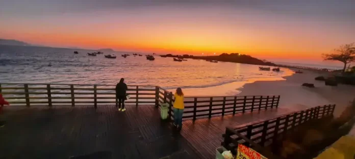 Pessoas observam o pôr do sol em um deque à beira-mar, com barcos ancorados e o céu alaranjado iluminando a praia.