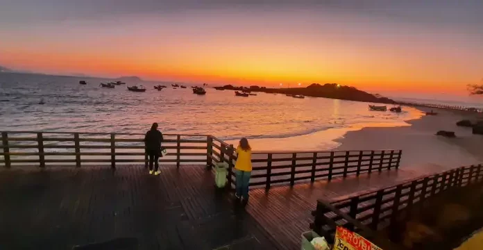 Pessoas observam o pôr do sol em um deque à beira-mar, com barcos ancorados e o céu alaranjado iluminando a praia.