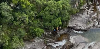 Imagem mostra bombeiros realizando o resgate de uma adolescente ferida em área de cachoeira, ilustrando a operação em terreno íngreme e de difícil acesso.