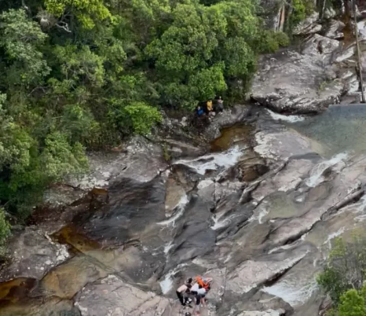 Imagem mostra bombeiros realizando o resgate de uma adolescente ferida em área de cachoeira, ilustrando a operação em terreno íngreme e de difícil acesso.