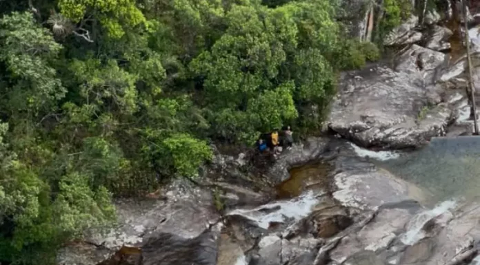Imagem mostra bombeiros realizando o resgate de uma adolescente ferida em área de cachoeira, ilustrando a operação em terreno íngreme e de difícil acesso.