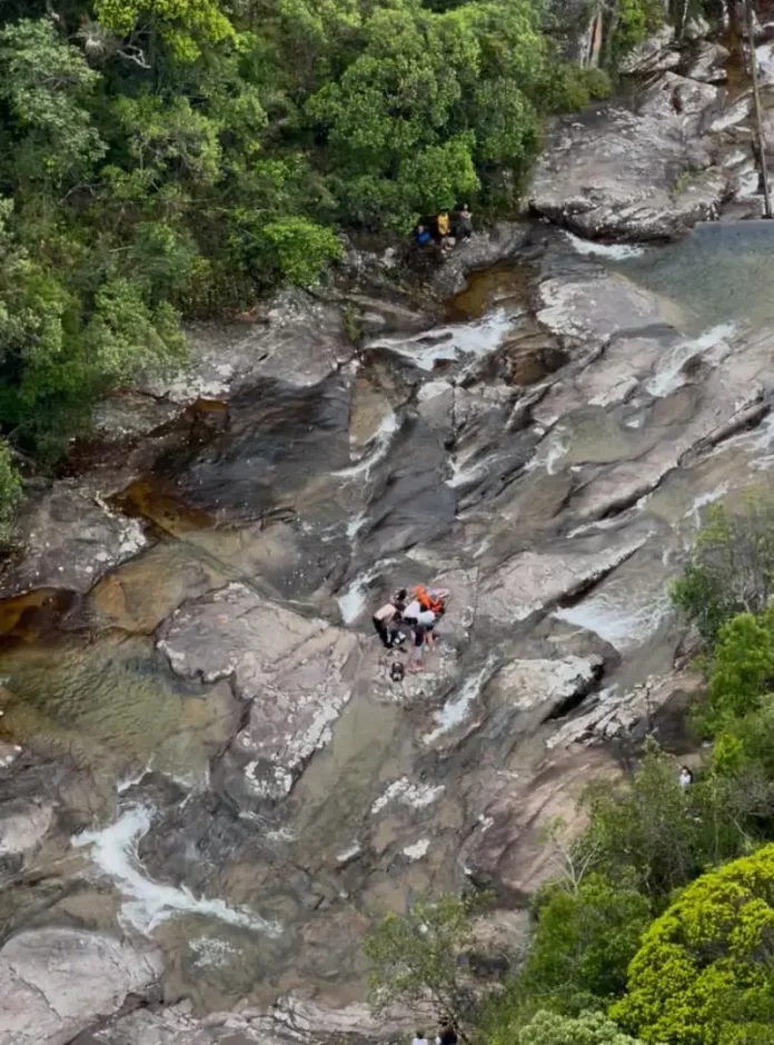 Imagem mostra bombeiros realizando o resgate de uma adolescente ferida em área de cachoeira, ilustrando a operação em terreno íngreme e de difícil acesso.