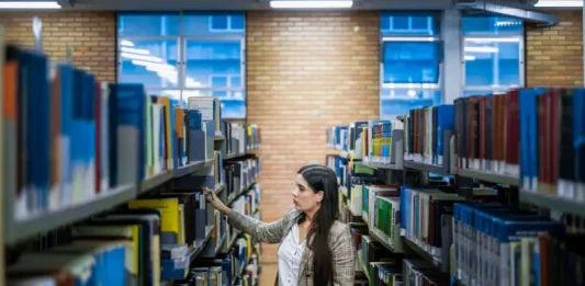 SC lança editais de renovação do Universidade Gratuita e do Fumdesc Aluna do Universidade Gratuita em uma biblioteca escolhendo livros, ilustrando o acesso ampliado à educação.