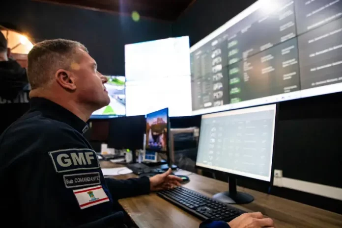 Agente da Guarda Municipal de Florianópolis monitora imagens da cidade em tempo real na central de videomonitoramento, demonstrando o uso de câmeras com tecnologia de reconhecimento facial para reforçar a segurança pública.