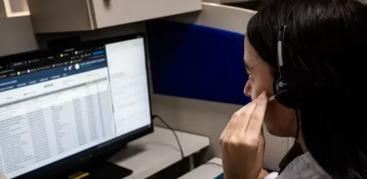 Atendente do Alô Saúde Floripa atende ligação de turista enquanto registra informações no computador, mostrando o serviço de suporte médico bilíngue da temporada.