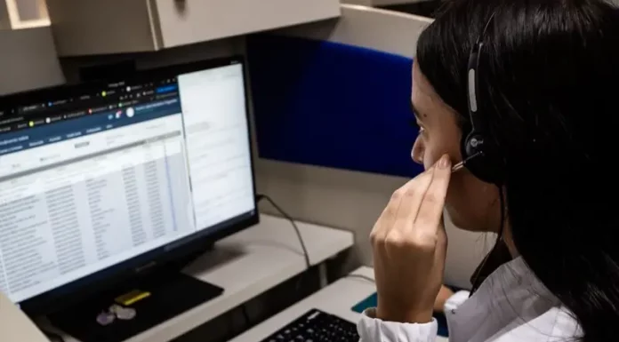 Atendente do Alô Saúde Floripa atende ligação de turista enquanto registra informações no computador, mostrando o serviço de suporte médico bilíngue da temporada.