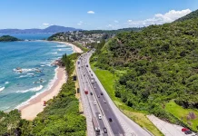 Operação Verão: Autopista Litoral Sul prevê 5,4 milhões de veículos entre Natal e Ano Novo Imagem aérea mostra trecho da BR-101 à beira-mar, com fluxo de veículos em rodovia cercada por vegetação e praia, ilustrando o movimento intenso previsto na Operação Verão.