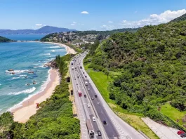 Imagem aérea mostra trecho da BR-101 à beira-mar, com fluxo de veículos em rodovia cercada por vegetação e praia, ilustrando o movimento intenso previsto na Operação Verão.